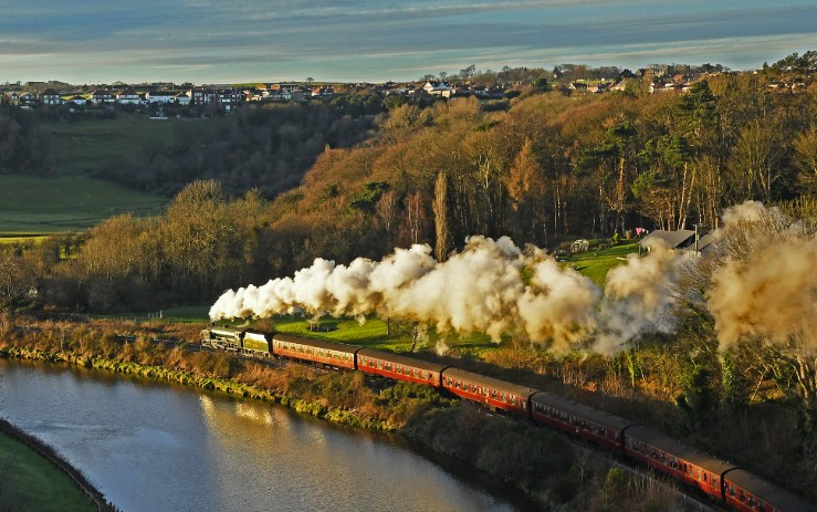 Steam train with red carriages running beside a river through wooded countryside
